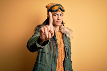 Young beautiful hispanic woman wearing ski glasses and coat for winter weather Pointing with finger up and angry expression, showing no gesture