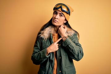 Young beautiful hispanic woman wearing ski glasses and coat for winter weather In hurry pointing to...