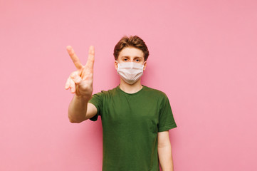 Young man in a protective medical mask stands on a pink background, looks at the camera and shows a...