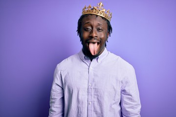 Young african american man wearing golden crown of king over isolated purple background sticking tongue out happy with funny expression. Emotion concept.