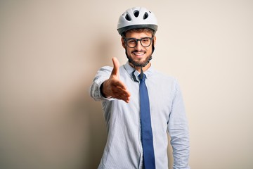 Young businessman wearing glasses and bike helmet standing over isolated white bakground smiling friendly offering handshake as greeting and welcoming. Successful business.