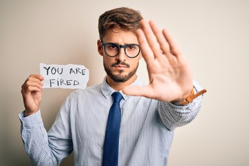 Young businessman wearing glasses holding paper with you are fired message with open hand doing stop sign with serious and confident expression, defense gesture