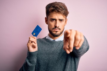Young customer man with beard holding credit card for payment over pink background pointing with...