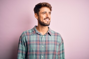 Young handsome man with beard wearing casual shirt standing over pink background looking away to...