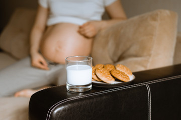 a pregnant woman's food; a pregnant woman is sitting in the background; glass of milk and a plate of oatmeal cookies in the background;