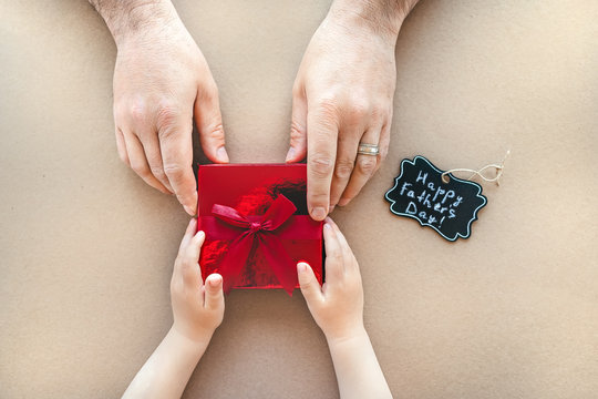 Happy Father's Day Concept. Son is giving a small red box with preset for his father. Flat lay, top view hands of man and boy - congratulation with present and black tag says Happy Father's Day!