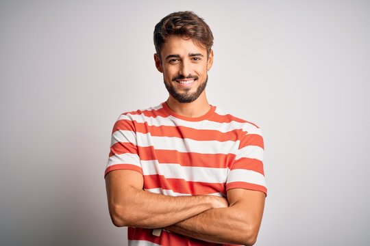 Young handsome man with beard wearing striped t-shirt standing over white background happy face smiling with crossed arms looking at the camera. Positive person.