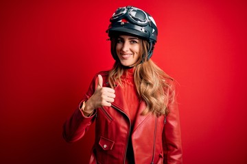 Young beautiful brunette motrocyclist woman wearing moto helmet over red background doing happy thumbs up gesture with hand. Approving expression looking at the camera showing success.