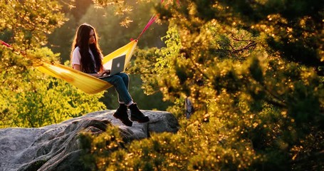 Awesome shot of hipster woman or millennial freelancer write blog post or work on remote office project on laptop. Girl sitting in hammock, relaxing on top of mountain