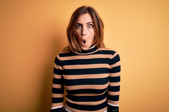 Young beautiful brunette woman wearing striped turtleneck sweater over yellow background afraid and shocked with surprise expression, fear and excited face.
