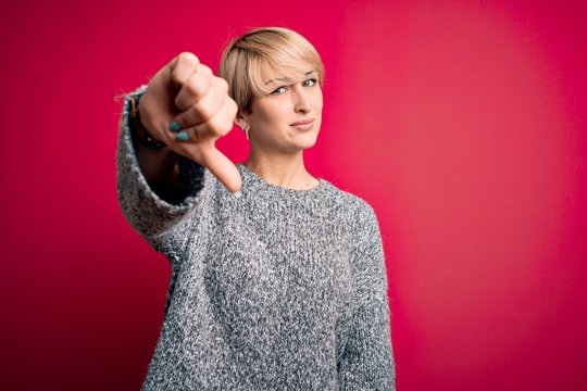 Young blonde woman with modern short hair wearing casual sweater over pink background looking unhappy and angry showing rejection and negative with thumbs down gesture. Bad expression.