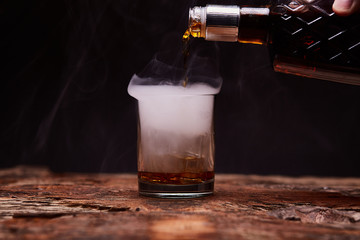 Glass of whiskey on wooden table. Pouring whiskey, smoke.