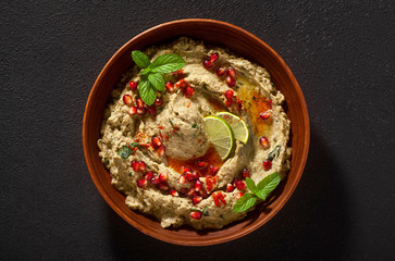 homemade spread of baked eggplant baba ganoush in a bowl with pomegranate seeds, lime, olive oil and lime slices on a black background