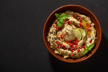 homemade spread of baked eggplant baba ganoush in a bowl with pomegranate seeds, lime, olive oil and lime slices on a black background