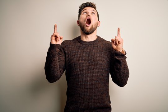 Young handsome blond man with beard and blue eyes wearing casual sweater standing amazed and surprised looking up and pointing with fingers and raised arms.