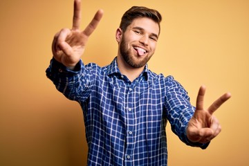 Young blond businessman with beard and blue eyes wearing shirt over yellow background smiling with tongue out showing fingers of both hands doing victory sign. Number two.