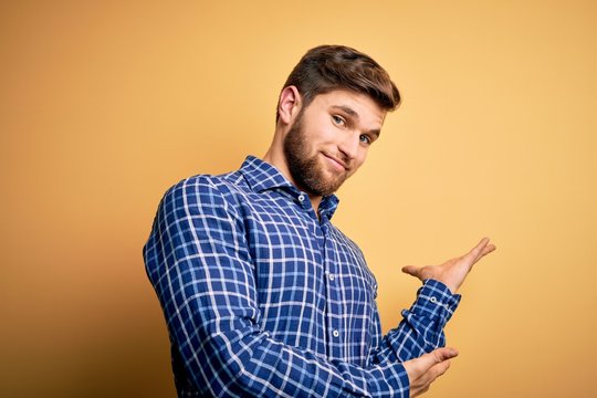 Young blond businessman with beard and blue eyes wearing shirt over yellow background Inviting to enter smiling natural with open hand