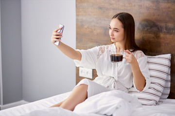 side view on charming woman taking photo on smartphone, she doing selfie with cup of coffee. she sits on bed at home, wearing white bathrobe