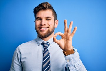 Young blond businessman with beard and blue eyes wearing elegant shirt and tie standing smiling positive doing ok sign with hand and fingers. Successful expression.