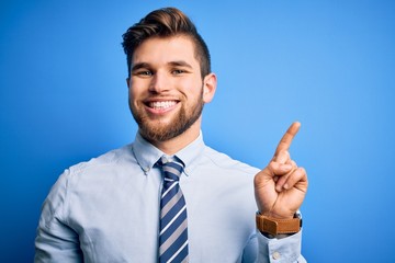Young blond businessman with beard and blue eyes wearing elegant shirt and tie standing showing and pointing up with finger number one while smiling confident and happy.