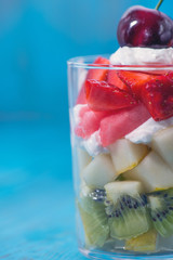 assorted diced fruits in a glass cup with cream and cherry on top as a decoration on a wooden blue background, vertical position