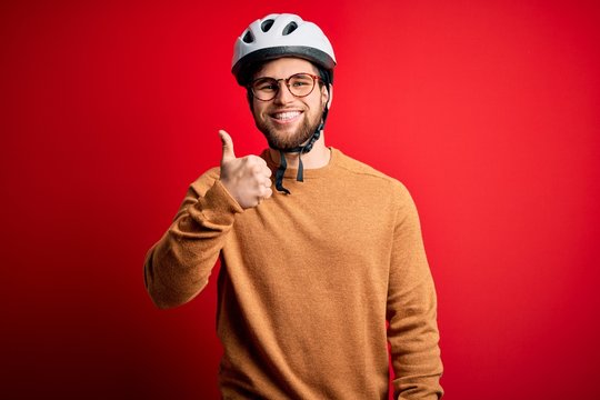 Young blond cyclist man with beard and blue eyes wearing bike helmet and glasses doing happy thumbs up gesture with hand. Approving expression looking at the camera showing success.