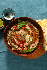 homemade spread of baked eggplant baba ganoush in a bowl with pomegranate seeds, lime, olive oil and lime slices on a blue linen tablecloth with flat bread