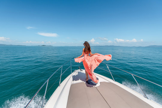 Outdoor shot of adorable young woman in a pink dress on edge of yacht, looking to beautiful nature landscape during trip. Happy woman enjoying summer travel. Back view