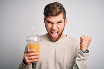 Fototapeta premium Young blond man with beard and blue eyes drinking healthy orange smoothie screaming proud and celebrating victory and success very excited, cheering emotion