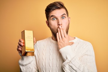 Young blond man with beard and blue eyes holding gold ingot over isolated yellow background cover...