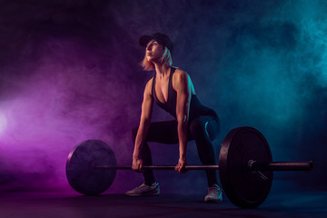 Fototapeta na wymiar Side view of female bodybuilder doing deadlift using barbell. Srtong woman with muscular body in sportswear training legs in dark atmosphere. Concept of bodybuilding.
