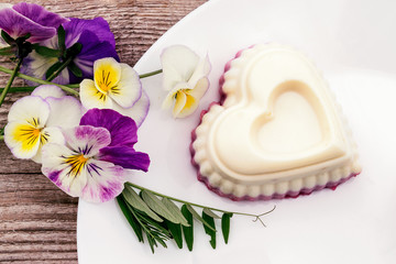 Fototapeta na wymiar Souffle in the shape of a heart with cottage cheese, agar-agar and cream on a white plate on a wooden background.Violet flowers adorn the composition.Сoncept of healthy food and healthy dessert