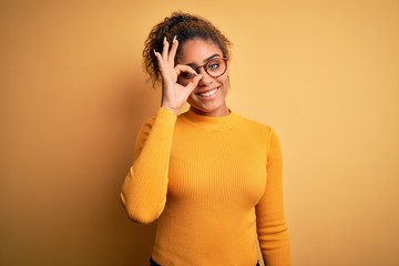 Young beautiful african american girl wearing sweater and glasses over yellow background doing ok gesture with hand smiling, eye looking through fingers with happy face.