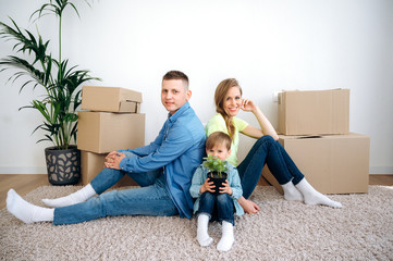 A beautiful happy family has moved to their new apartment. They are sitting on the floor among cardboard boxes in casual clothes and smiling, the little boy is holding small flowerpot in his hands