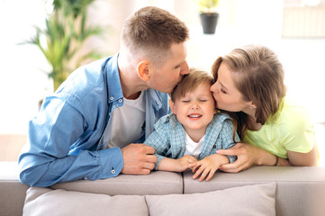 Fototapeta premium Portrait of a happy family. Dad and mom gently kiss their little beloved son on a sofa at home. Parents and little son