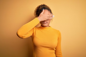 Young beautiful african american girl wearing sweater and glasses over yellow background covering eyes with hand, looking serious and sad. Sightless, hiding and rejection concept