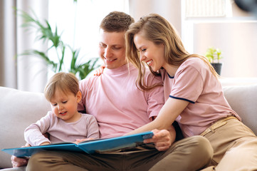 Obraz premium Family leisure at home. Young happy parents read a children's book with their adorable little son, sitting at home on the couch