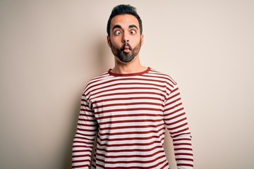 Young handsome man with beard wearing casual striped t-shirt standing over white background making fish face with lips, crazy and comical gesture. Funny expression.