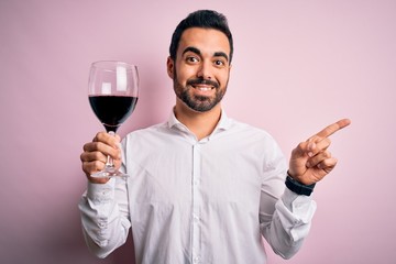 Young handsome man with beard drinking glass of red wine over isolated pink background very happy pointing with hand and finger to the side