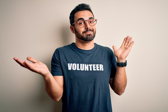 Handsome man with beard wearing t-shirt with volunteer message over white background clueless and confused expression with arms and hands raised. Doubt concept.