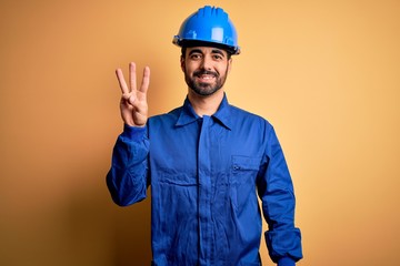 Mechanic man with beard wearing blue uniform and safety helmet over yellow background showing and pointing up with fingers number three while smiling confident and happy.