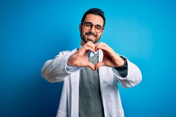 Young handsome doctor man with beard wearing coat and glasses over blue background smiling in love doing heart symbol shape with hands. Romantic concept.