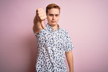 Young handsome redhead man wearing casual summer shirt standing over pink background looking unhappy and angry showing rejection and negative with thumbs down gesture. Bad expression.