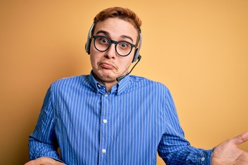 Fototapeta premium Young handsome redhead call center agent man wearing glasses working using headset clueless and confused expression with arms and hands raised. Doubt concept.