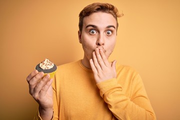 Young handsome redhead man eating sweet chocolate cupcake dessert over yellow background covering...
