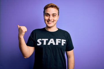 Young handsome redhead worker man wearing staff t-shirt uniform over purple background Pointing to the back behind with hand and thumbs up, smiling confident