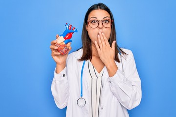 Young beautiful brunette cardiologist woman wearing stethoscope holding plastic heart covering...