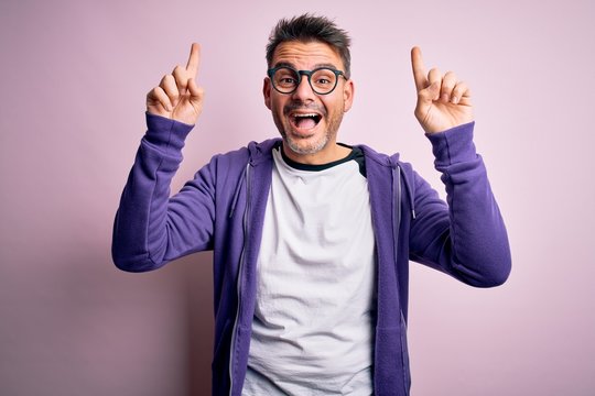 Young handsome man wearing purple sweatshirt and glasses standing over pink background smiling amazed and surprised and pointing up with fingers and raised arms.