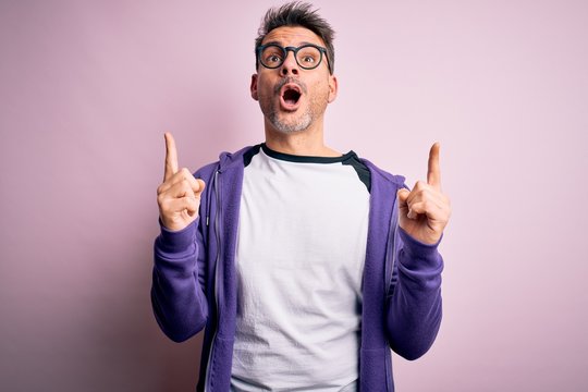 Young handsome man wearing purple sweatshirt and glasses standing over pink background amazed and surprised looking up and pointing with fingers and raised arms.