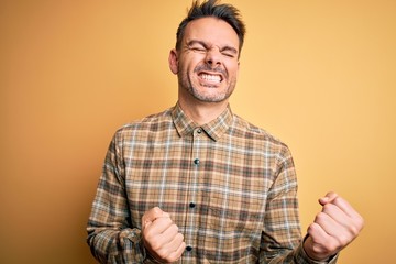 Young handsome man wearing casual shirt standing over isolated yellow background very happy and excited doing winner gesture with arms raised, smiling and screaming for success. Celebration concept.